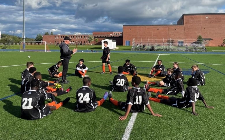 A group of soccer players sat on a field in a circle with a man stood in the circle talking.