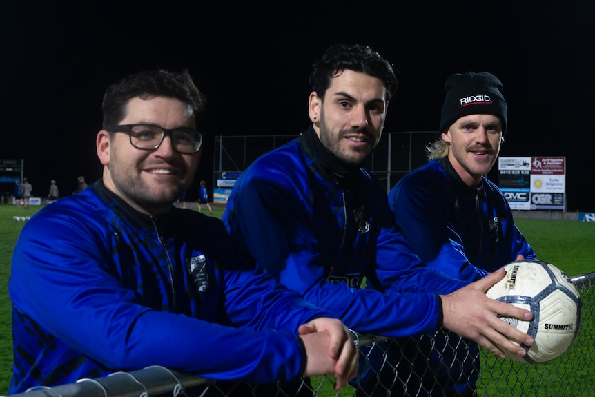 Three male soccer players wearing blue and black leaning on a fence on the side of a soccer ground
