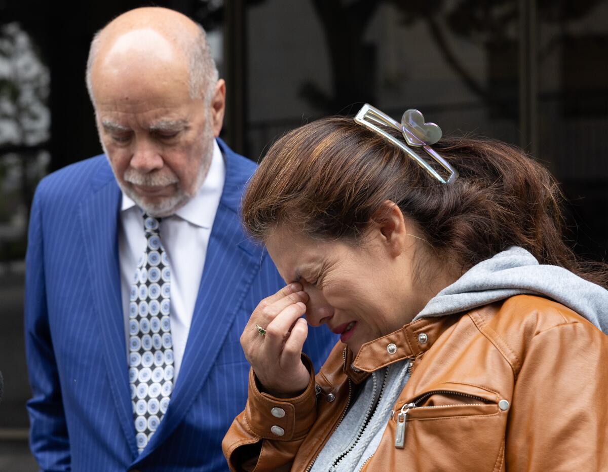 A woman with brown hair, in a hoodie and brown jacket, holds fingers to her nose. Next to her is a man in a dark blue suit